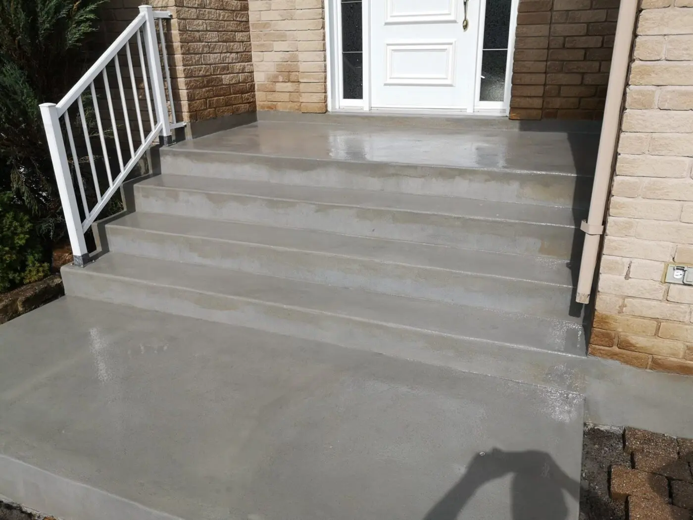 A freshly poured concrete staircase with five steps leads up to a white door on a brick building in Plantation, FL. The steps have a smooth, wet appearance, and a white metal handrail is on the left side. The shadow of someone taking the photo is visible at the bottom of the image. For expert concrete contracting services and a free quote, look no further!