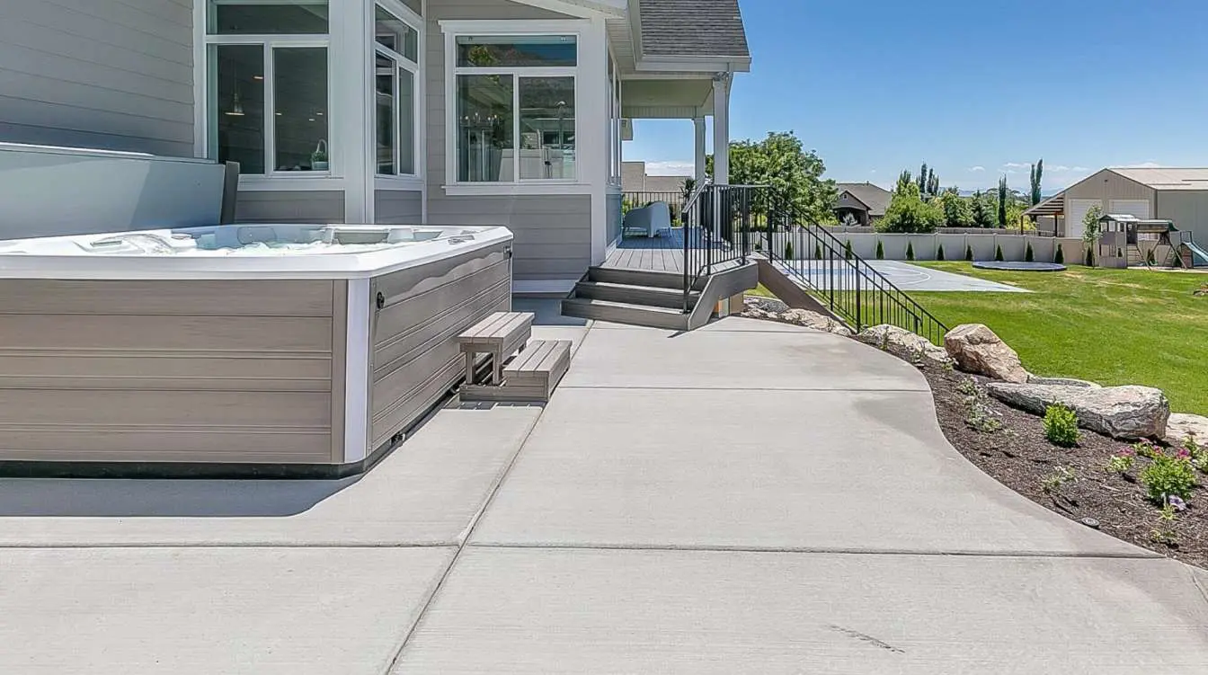 A spacious backyard in Plantation, FL features a modern hot tub with small steps on a concrete patio. The area includes a well-manicured lawn, a garden bed with flowers, and views of neighboring houses and trees. Steps and a railing lead to the house entrance. Contact us for concrete contracting services or a free quote!