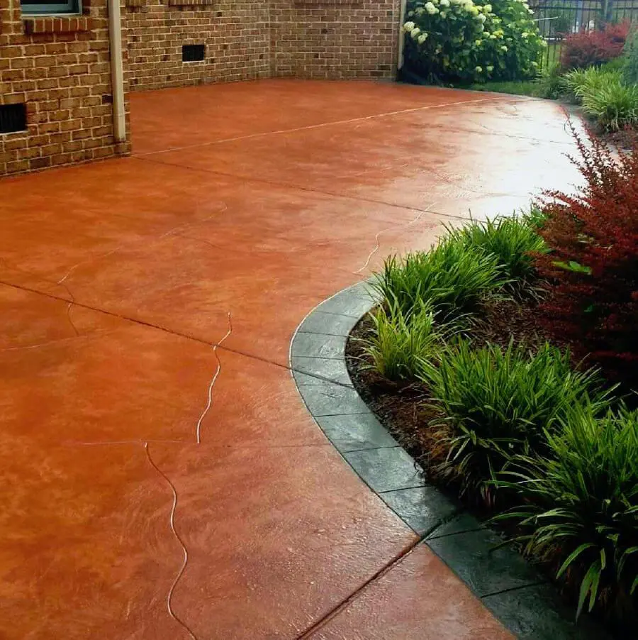 A long driveway with a freshly washed and finished concrete surface, courtesy of top-tier Concrete Contractors Miami, leads to a residential garage. The driveway is bordered by red bricks and surrounded by garden beds and neatly trimmed bushes. A garden hose lays on the left, with wrapped objects on the right side.